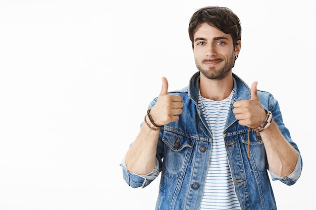 Foto gratuita ritratto di un cliente maschio di bell'aspetto soddisfatto con occhi azzurri e setola in giacca di jeans che mostra i pollici in su e sorride soddisfatto, esprimendo un'opinione positiva sul prodotto