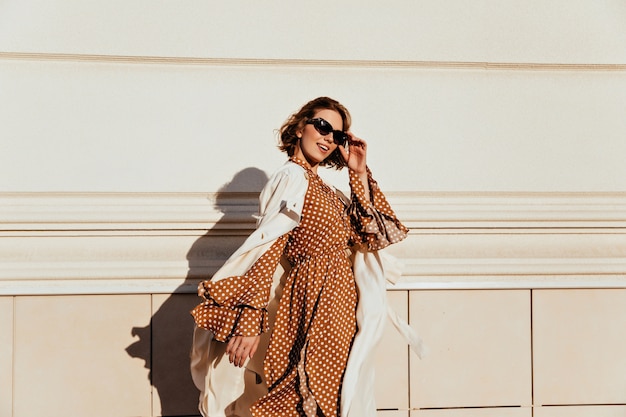 Foto gratuita bella donna in abito lungo marrone godendo la giornata di sole. amabile ragazza bianca in abbigliamento retrò che cammina per strada.