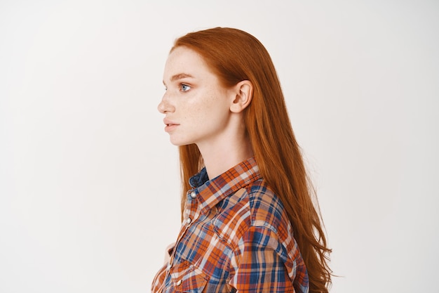 Foto gratuita profilo di una giovane studentessa dai capelli rossi con lunghi capelli naturali allo zenzero e pelle pallida, guardando a sinistra, in piedi in camicia casual sul muro bianco