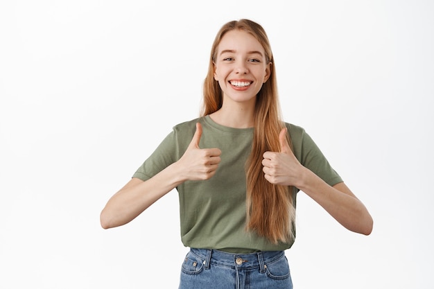 Foto gratuita una bella studentessa sorridente mostra i pollici in su, sembra soddisfatta e annuisce in segno di approvazione, darle supporto, consigliare il prodotto, elogiare la qualità eccellente, in piedi sul muro bianco