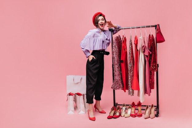 Foto gratuita spettacolare signora in pantaloni neri solleva civettuola la gamba e tiene la borsa su sfondo rosa con i pacchetti dopo lo shopping.