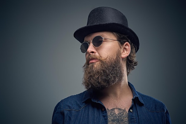 Foto gratuita ritratto in studio di un elegante maschio barbuto con un tatuaggio sul petto, vestito con una camicia di jeans, un cappello a cilindro e occhiali da sole isolati su sfondo grigio.