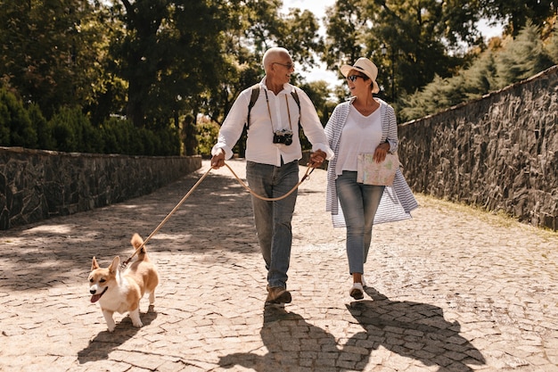 Foto gratuita donna alla moda in cappello e camicetta blu che cammina e che guarda uomo dai capelli grigi in camicia bianca a maniche lunghe con fotocamera e corgi nel parco.