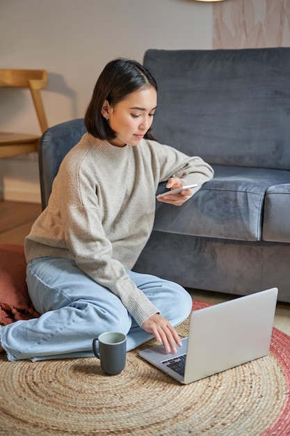 Foto gratuita ripresa verticale di una giovane donna in una casa accogliente che lavora al computer portatile utilizzando lo smartphone e bevendo caffè seduti