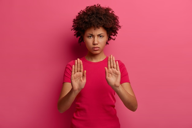 Foto gratuita la vita in su di una donna seria dalla pelle scura mostra abbastanza o nessun gesto, guarda con rabbia, tira i palmi verso, rifiuta o disapprova qualcosa di brutto, dice fermarsi qui proibisce l'azione