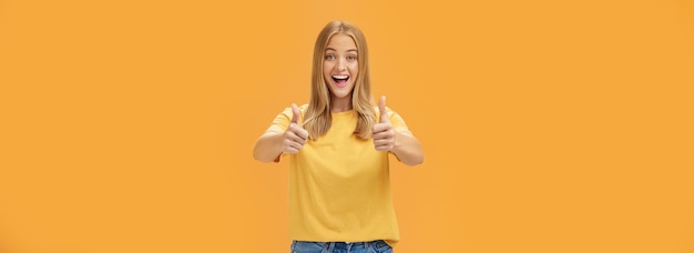 Foto gratuita la donna sostiene con i pollici alzati e divertito sorriso allegro mostrando atteggiamento positivo esprimendo come su concetto o idea dando approvazione posando felice e deliziato contro uno sfondo arancione