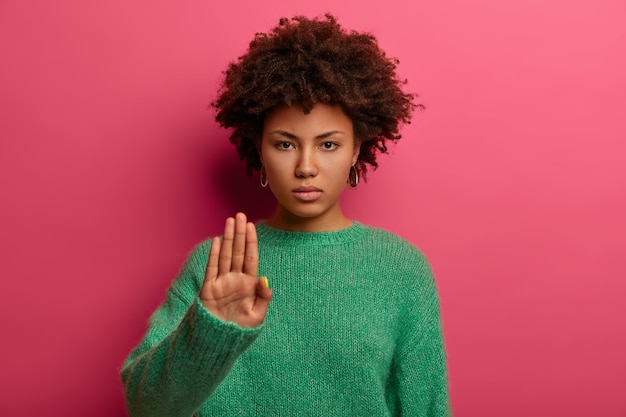 Foto gratuita la giovane donna afroamericana dai capelli ricci seria fa il segnale di stop con il palmo, indossa un maglione verde, dimostra divieto e restrizione, rifiuta qualcosa, modella contro il muro rosa, dice di no