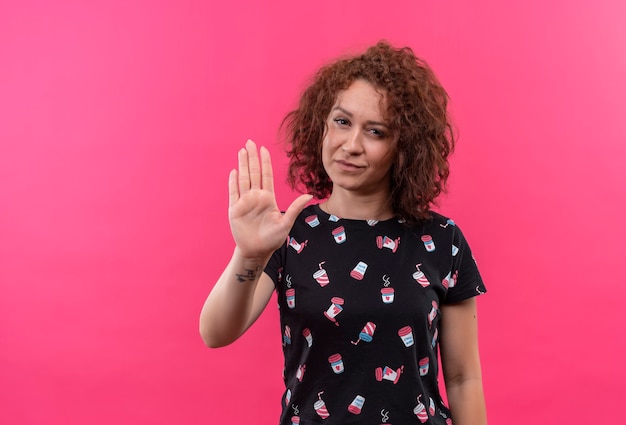 Foto gratuita giovane donna con i capelli ricci corti che fa il fanale di arresto con la mano che osserva seriamente che sta sopra la parete rosa