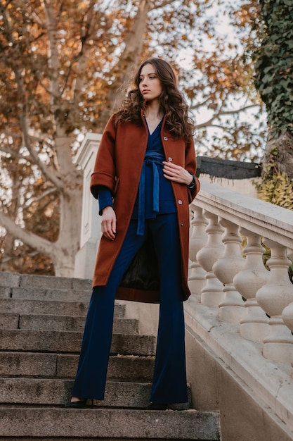 Photo gratuite belle élégante femme maigre souriante aux cheveux bouclés marchant dans les escaliers de la rue vêtue d'un manteau brun chaud et d'un costume bleu, style de rue à la mode automne