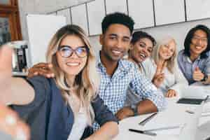 Photo gratuite employé de bureau noir en chemise à carreaux embrassant une femme secrétaire blonde pendant qu'elle fait selfie. jeunes managers d'entreprise internationale s'amusant lors d'une réunion.