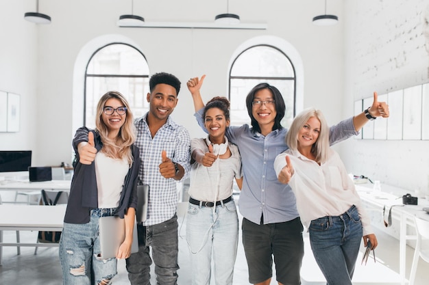 Photo gratuite Étudiante blonde en jeans déchirés tenant un ordinateur portable et riant après un test difficile. un type asiatique dans des verres agitant les mains, célébrant la fin des examens et le début des vacances.