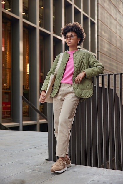 Photo gratuite femme aux cheveux bouclés concentré vers l'avant avec une expression confiante porte des lunettes de soleil veste pantalon baskets attend que quelqu'un se promène en ville