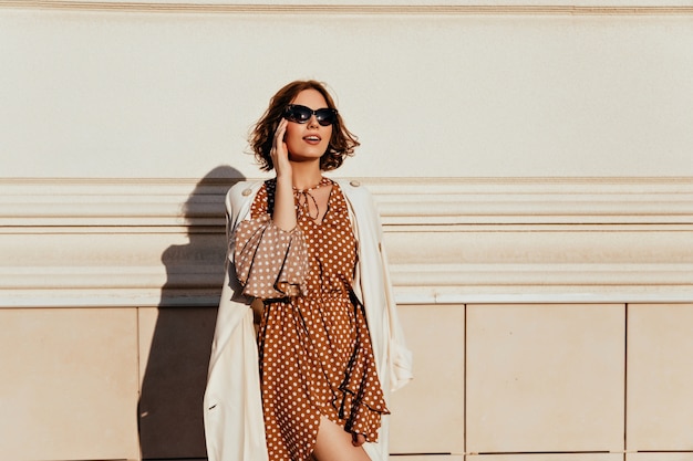 Photo gratuite femme galbée en robe vintage touchant ses lunettes. prise de vue en extérieur d'une fille détendue intéressée en tenue brune.