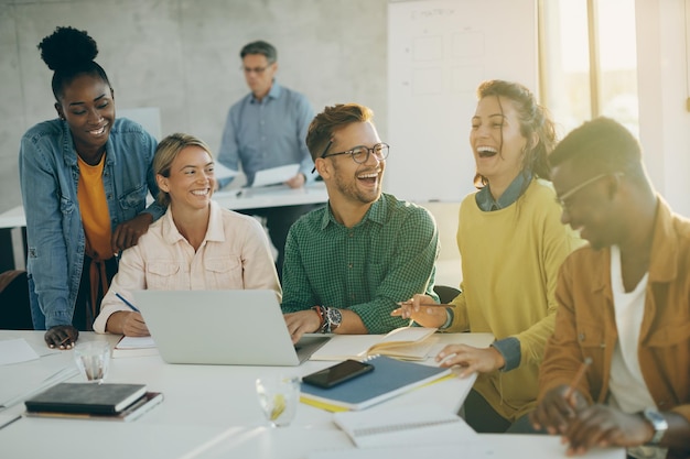 Photo gratuite groupe d'étudiants heureux s'amusant et riant tout en utilisant un ordinateur portable et en étudiant en classe. leur professeur est en arrière-plan.