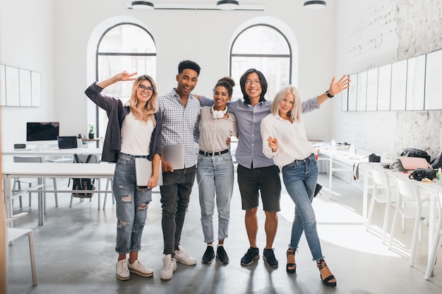 Photo gratuite portrait en pied de femme mince employé de bureau en jeans debout avec les jambes croisées près d'un collègue asiatique. photo intérieure d'un grand étudiant africain et d'une femme européenne heureuse avec un ordinateur portable.