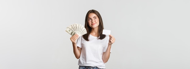 Photo gratuite réfléchie fille souriante tenant de l'argent et une carte de crédit regardant le coin supérieur gauche debout sur fond blanc et réfléchissant