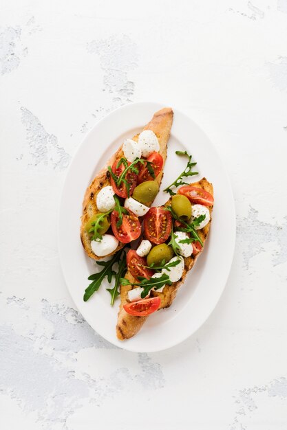 Foto bruschetta caprese toast con pomodorini, mozzarella, olive e basilico sulla vecchia superficie chiara del piatto bianco. vista dall'alto.