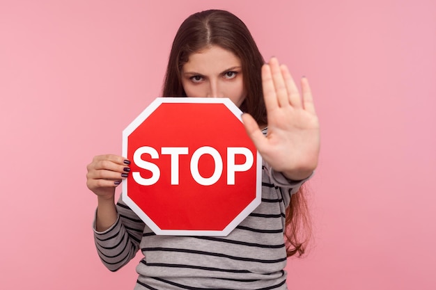 Foto nessun ritratto proibito di una ragazza severa e prepotente che guarda con rabbia e mostra un gesto di arresto tenendo il segnale stradale come avvertimento di divieto di accesso vietato in studio indoor girato isolato su sfondo rosa