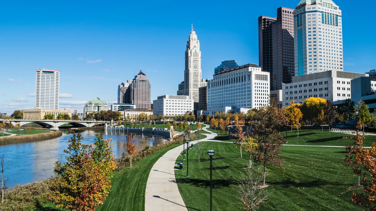 A daytime shot of Columbus, Ohio, with a bridge, river and green space in the foreground. Tall buildings dot the background.