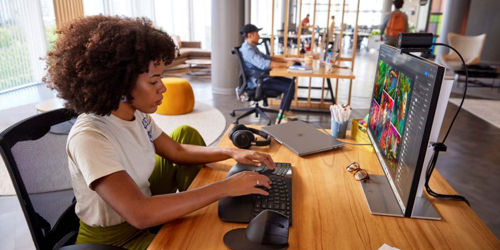 A woman with curly hair works at an HP desktop computer in a modern open office, editing a colorful image on a large monitor with other colleagues visible in the background.