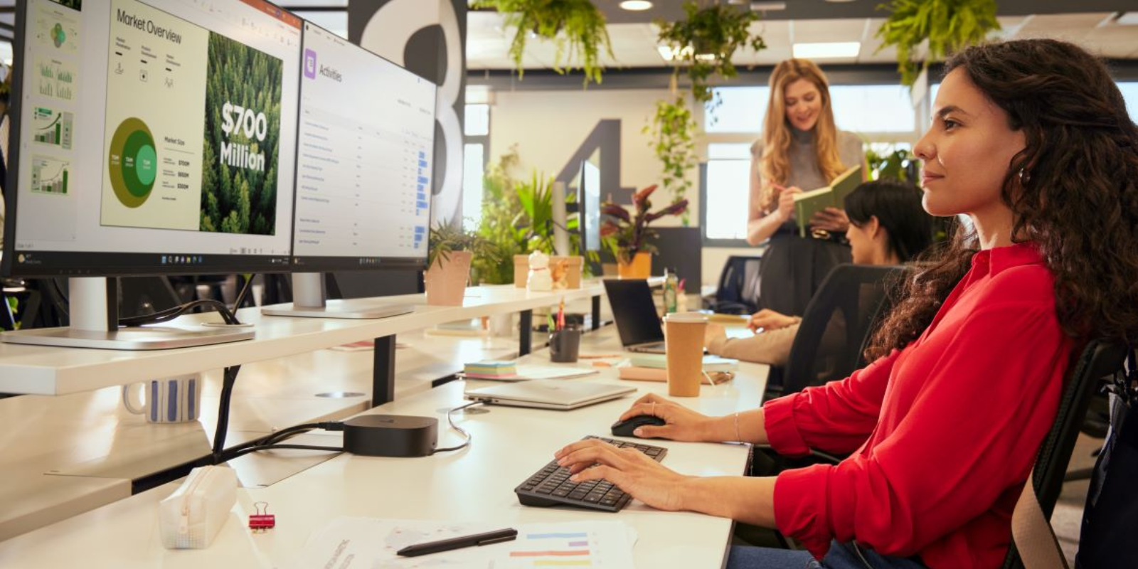 Modern office with woman in red top working at computer displaying market data. Colleagues in background. Plants hanging from ceiling in an open workspace. 