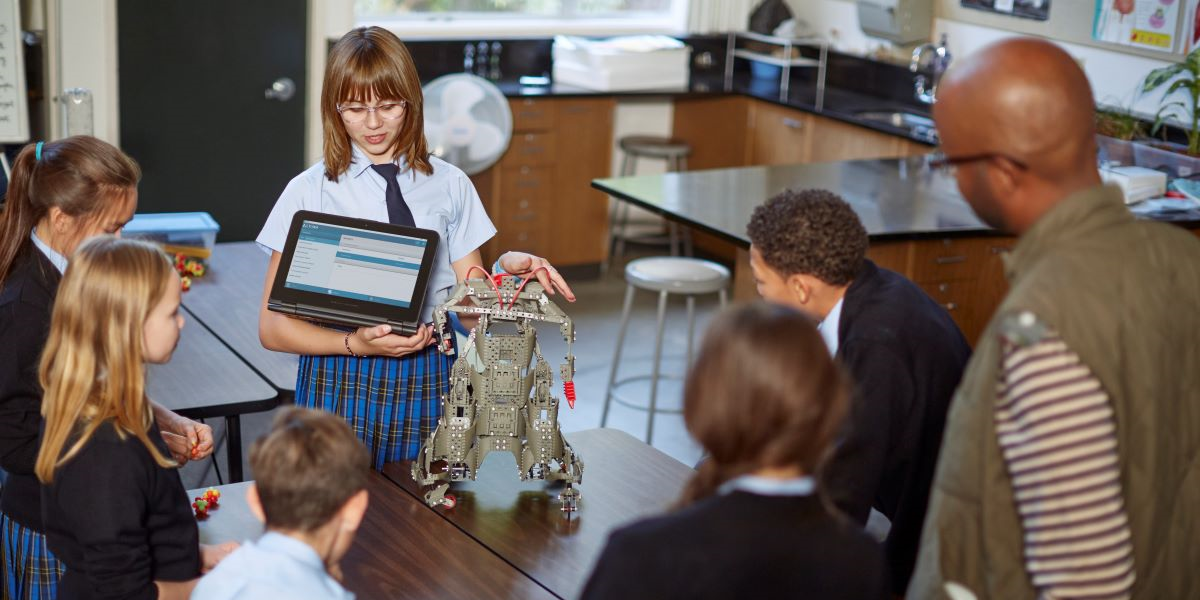 Student presenting robot model to classmates and teacher in science lab. Girl holds a tablet and demonstrates metal structure. Diverse group attentively watching.