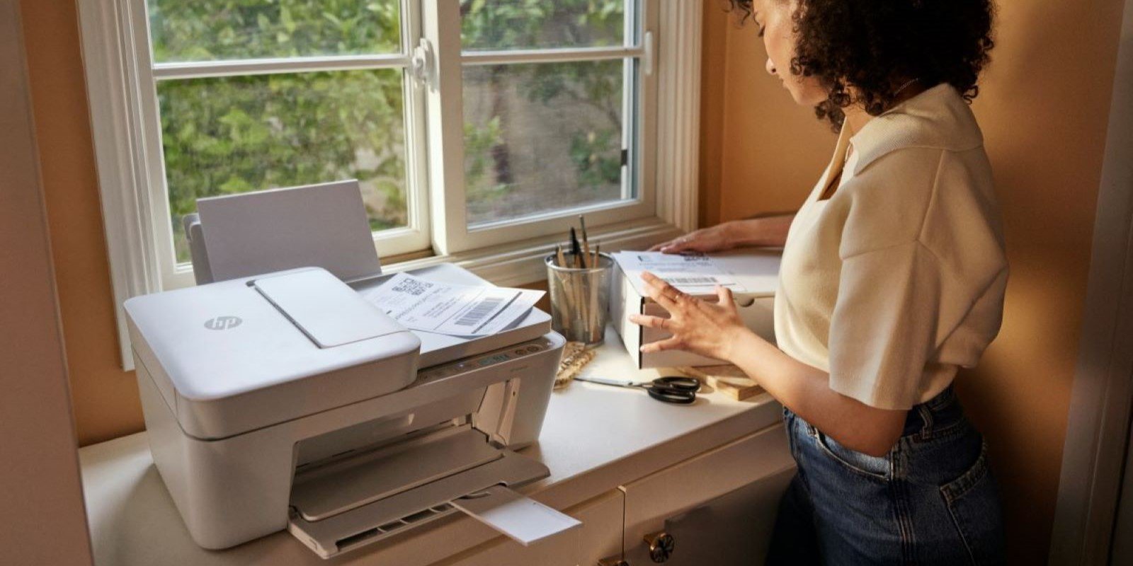 Woman with curly hair examines documents next to HP printer on desk by window. Sunlit room with view of trees outside. Office supplies visible on desk