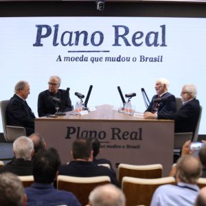 Foto de uma bancada com quatro homens brancos sentados conversando. Ao fundo, um painel escrito Plano Real, a moeda que mudou o Brasil.