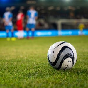 Imagem de uma bola de futebol em campo. Ao fundo atletas e a arquibancada.