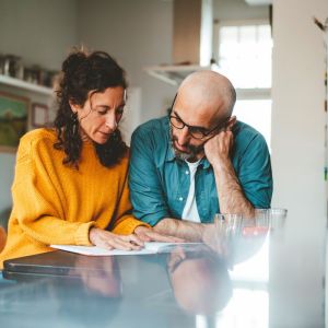 Foto de um casal olhando para uma folha em papel. A mulher é morena e veste uma blusa de frio laranja. O homem é careca e veste uma camisa azul.