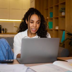 Imagem de uma mulher negra, em uma sala sentada em frente a um notebook. Em cima da mesa tem uma calculadora e apostilas.