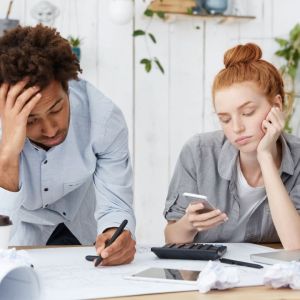 Imagem de um homem negro com uma das mãos na cabeça, escrevendo em um papel. Ao seu lado, uma mulher branca, olhando para o celular. Em cima da mesa, vários papeis e uma calculadora.