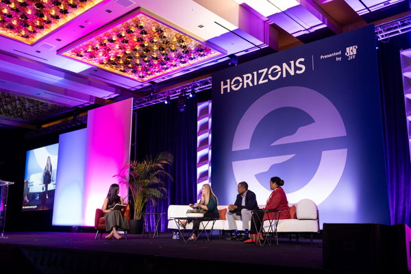 A panel of four people seated on stage at the Horizons conference, discussing under colorful lighting. A large screen and decorative plants are in the background.