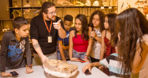 Um grupo de crianças junto a um monitor do Museu de Zoologia da USP que explica sobre um fóssil da fauna brasileira exposto numa mesa