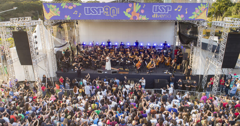 Palco em área aberta com músicos de orquestra e o público vendo a apresentação em pé