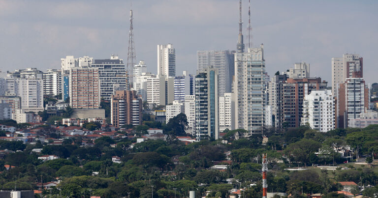 Vista de uma grande cidade mostrando uma área com árvores na parte de baixo e prédios e torres na parte cima. O céu está nublado