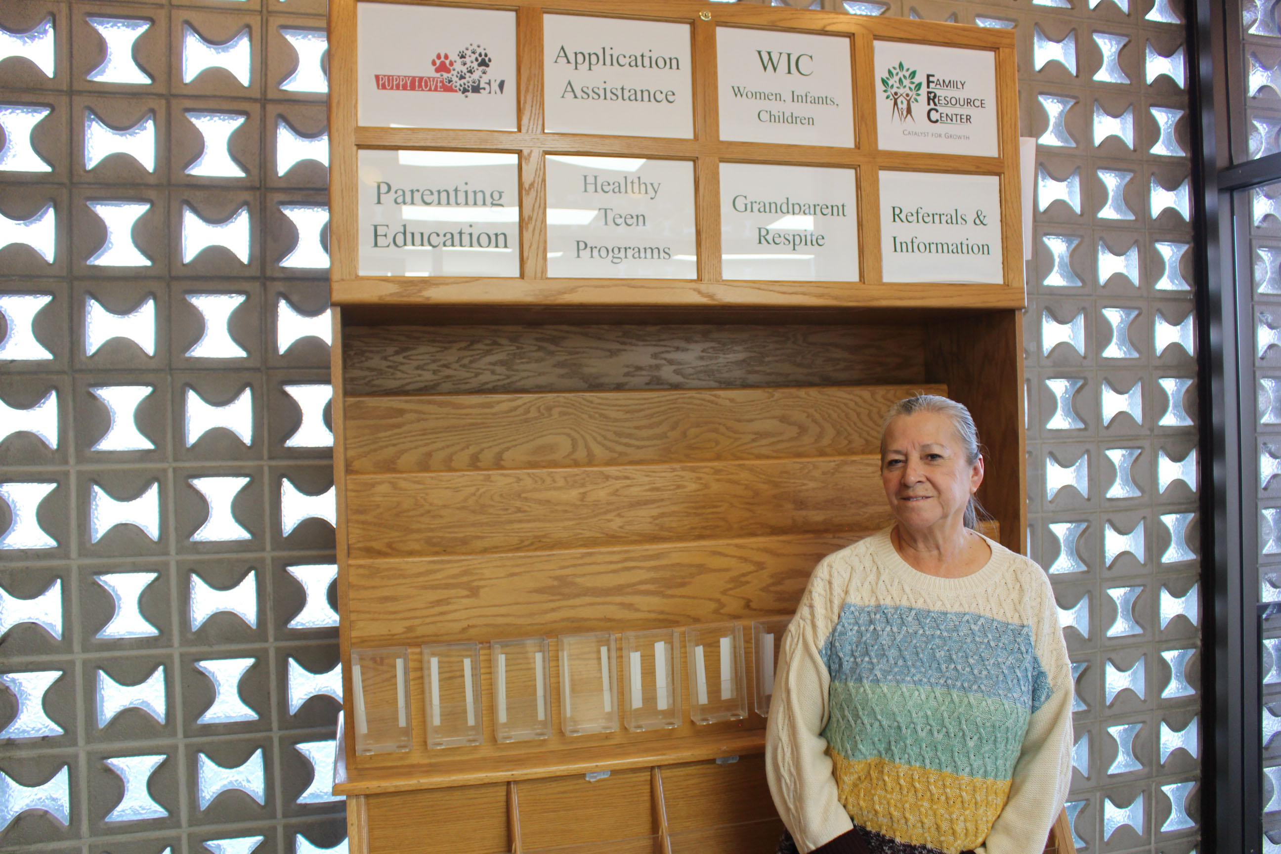 A portrait of a woman standing beside a wall and smiling. On the wall, signs read: "Puppy Love P5K / Application Assistance / WIC – Women, Infants, Children / Family Resource Center / Parenting Education / Healthy Teen Programs / Grandparent Respite / Referrals & Information"