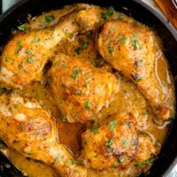 A cast-iron skillet resting on a tea towel. Five pieces of bone-in chicken in gravy are in the skillet.