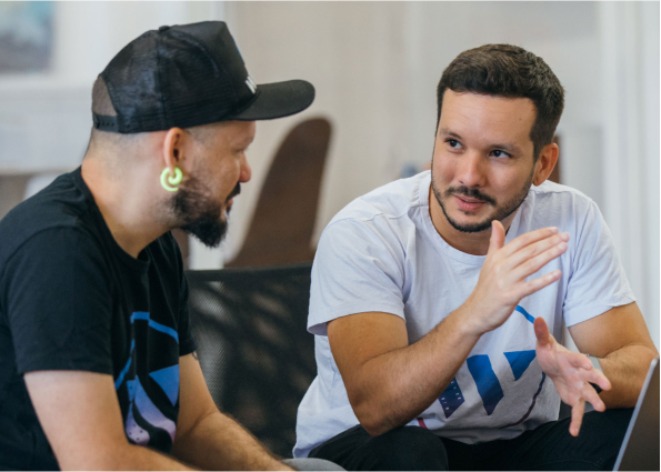 A man in focus is discussing with other man wearing black cap while sitting in front of laptop.