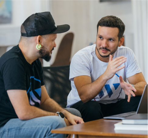 A man in focus is discussing with other man wearing black cap while sitting in front of laptop.