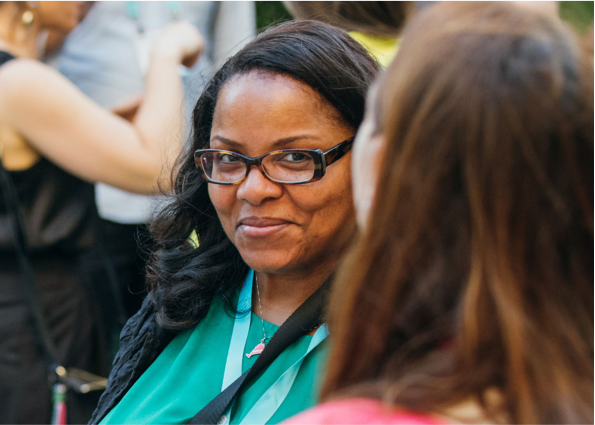 A dark woman with long hair and wearing spectacles smiling at other woman in crowd.