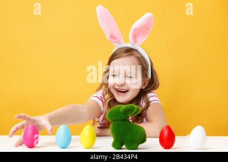 Easter concept. Happy kid in bunny ears yellow background. A little girl sits at a table and hunts for colored eggs. Stock Photo