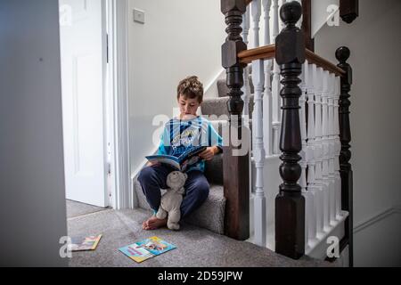 Six year old boy reading a book whilst sat on his residential home staircase, England, United Kingdom Stock Photo