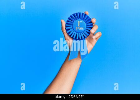 Hand of hispanic man holding 1st place ribbon over isolated blue background. Stock Photo