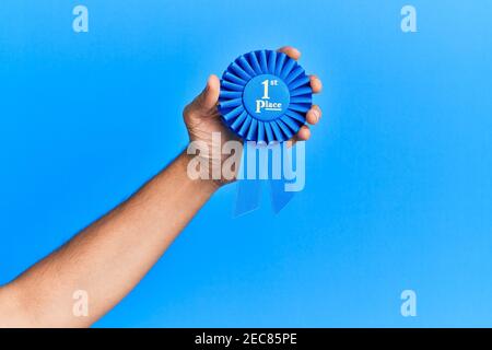 Hand of hispanic man holding 1st place ribbon over isolated blue background. Stock Photo