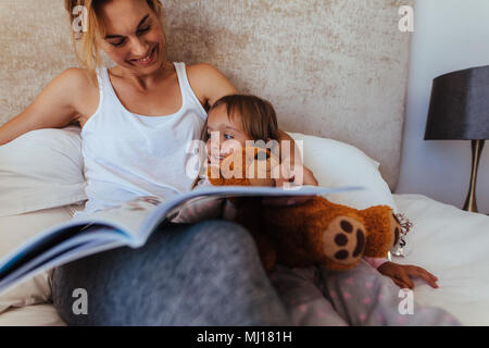 Happy family reading bedtime story in bed. Woman looking at her daughter and smiling while reading a book in bedroom. Stock Photo