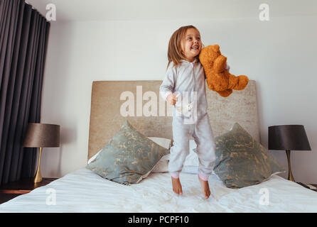 Joyful little girl jumping on bed with teddy bear. Child girl playing on the bed in her bedroom and smiling. Stock Photo