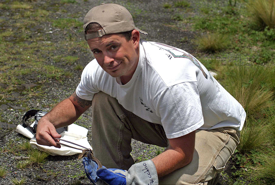 A man squats on scrubby ground while holding a tiny snake with tongs and thick, leather gloves.