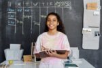 A student holds her notebook and pen while standing in front of her chalkboard at home with her study schedule written on it.