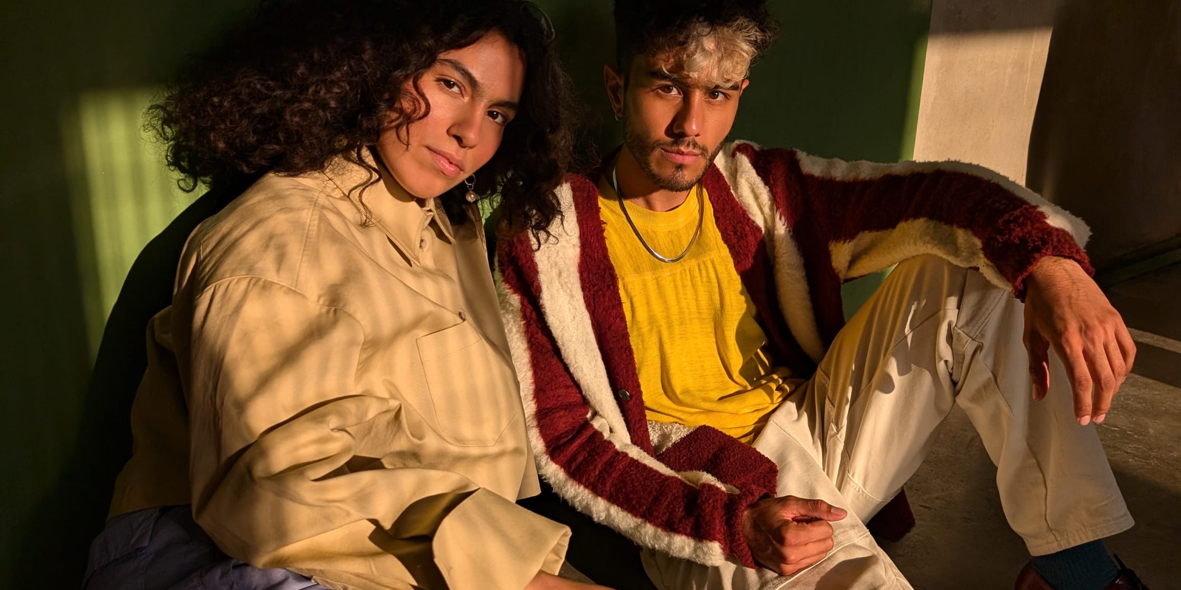 Two people pose seated against a wall in dim light.